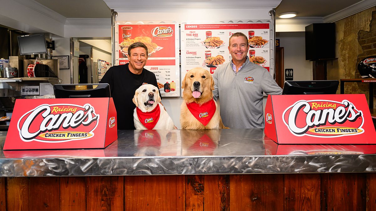 Todd Graves and Kirk Herbstreit stand behind a Raising Cane's counter with two dogs in Cane's bandanas.