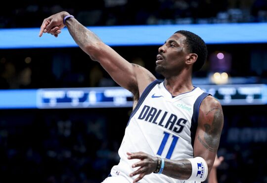 Dallas Mavericks guard Kyrie Irving holds his follow-through after a shot in a white Dallas jersey.