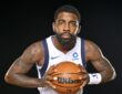 Dallas Mavericks guard Kyrie Irving leans toward the camera gripping a basketball with both hands against a black backdrop.
