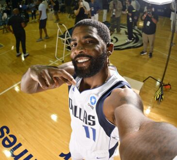 Dallas Mavericks guard Kyrie Irving smiles and points at himself while holding the camera at arm's length on the practice court.