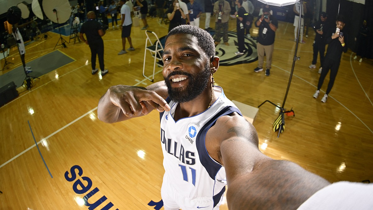 Dallas Mavericks guard Kyrie Irving smiles and points at himself while holding the camera at arm's length on the practice court.