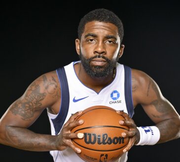 Dallas Mavericks guard Kyrie Irving leans toward the camera gripping a basketball with both hands against a black backdrop.