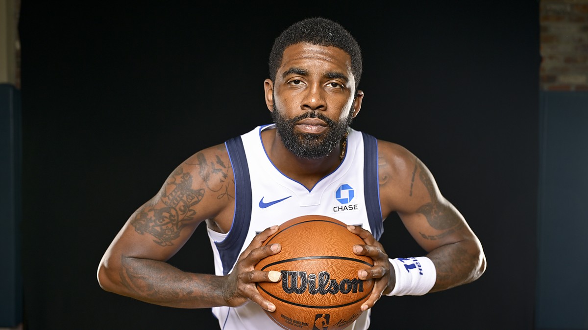 Dallas Mavericks guard Kyrie Irving leans toward the camera gripping a basketball with both hands against a black backdrop.