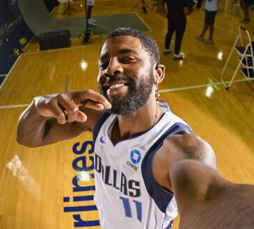 Dallas Mavericks guard Kyrie Irving grins while holding the camera at arm's length for a selfie on a gym floor.