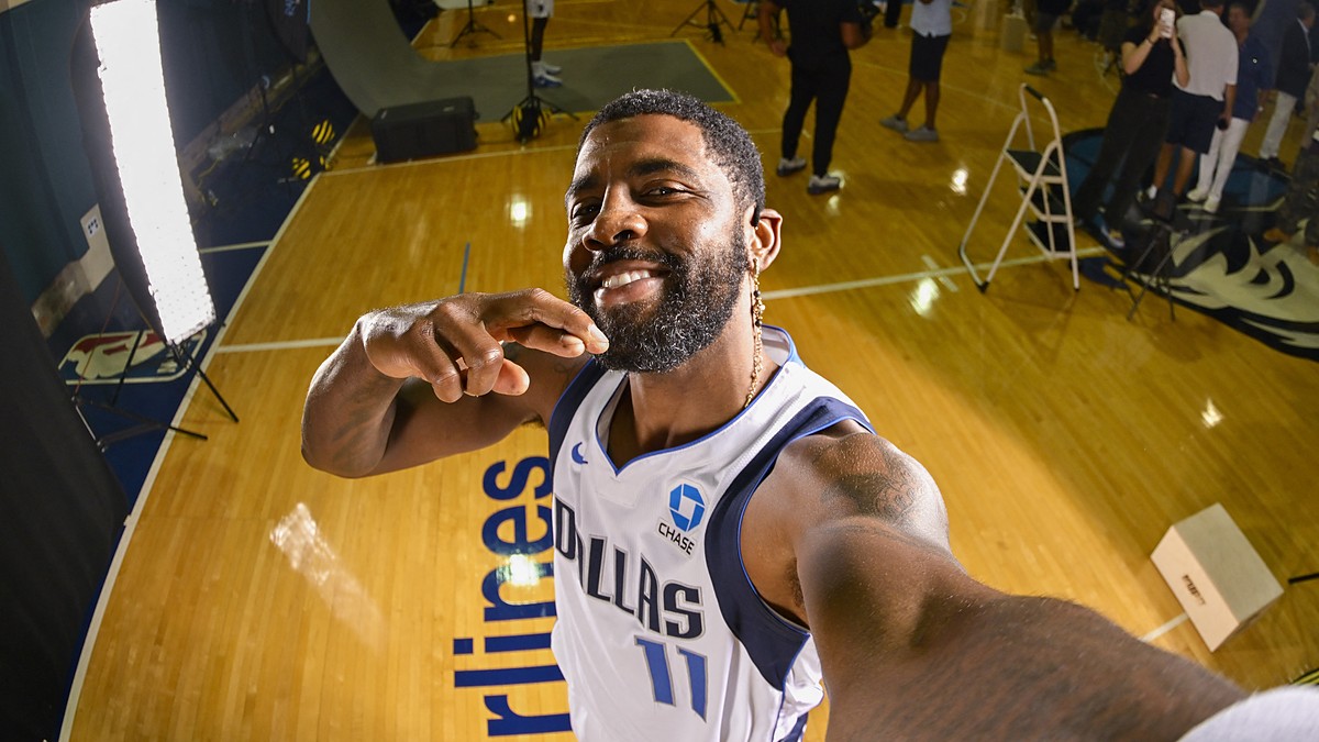 Dallas Mavericks guard Kyrie Irving grins while holding the camera at arm's length for a selfie on a gym floor.