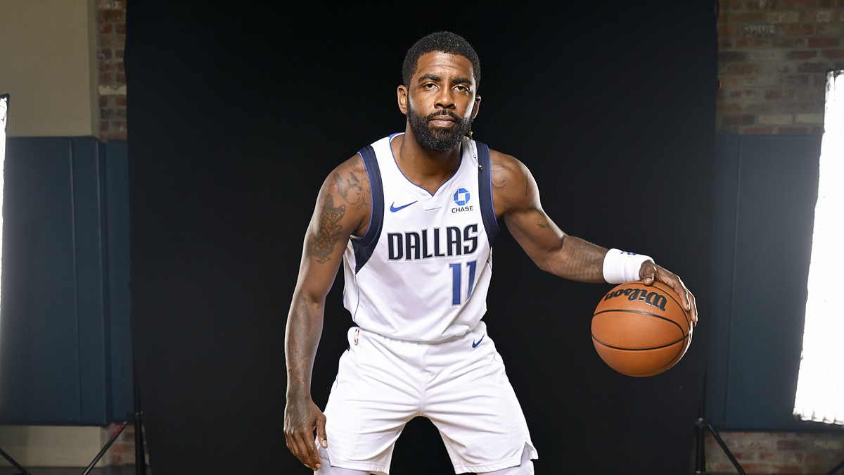 Dallas Mavericks guard Kyrie Irving crouches in a dribbling stance with a basketball against a black backdrop.