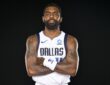 Dallas Mavericks guard Kyrie Irving stands with his arms crossed in a white Dallas jersey against a black backdrop.