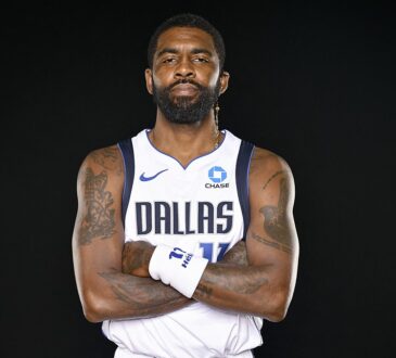 Dallas Mavericks guard Kyrie Irving stands with his arms crossed in a white Dallas jersey against a black backdrop.