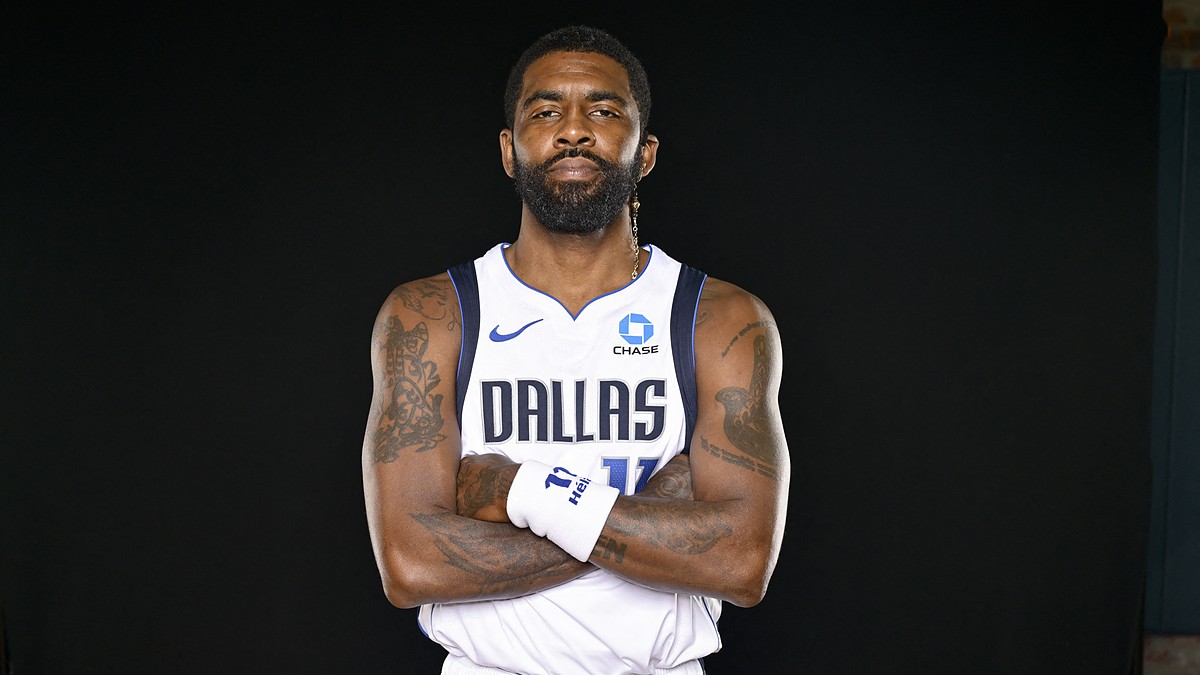 Dallas Mavericks guard Kyrie Irving stands with his arms crossed in a white Dallas jersey against a black backdrop.
