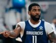 Dallas Mavericks guard Kyrie Irving points to his right while wearing a blue No. 11 practice jersey over a white shirt.