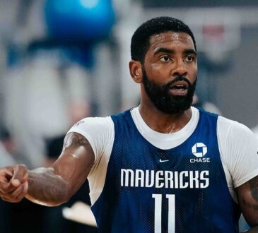 Dallas Mavericks guard Kyrie Irving points to his right while wearing a blue No. 11 practice jersey over a white shirt.