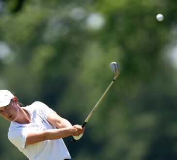 Golfer Lindy Duncan follows through on an iron shot as the ball flies against a backdrop of blurred trees.
