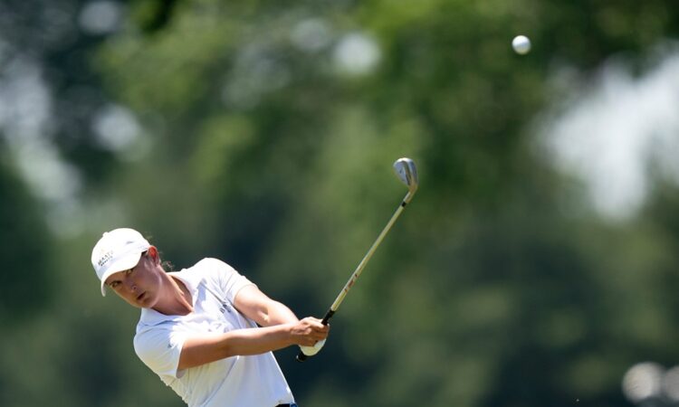 Golfer Lindy Duncan follows through on an iron shot as the ball flies against a backdrop of blurred trees.
