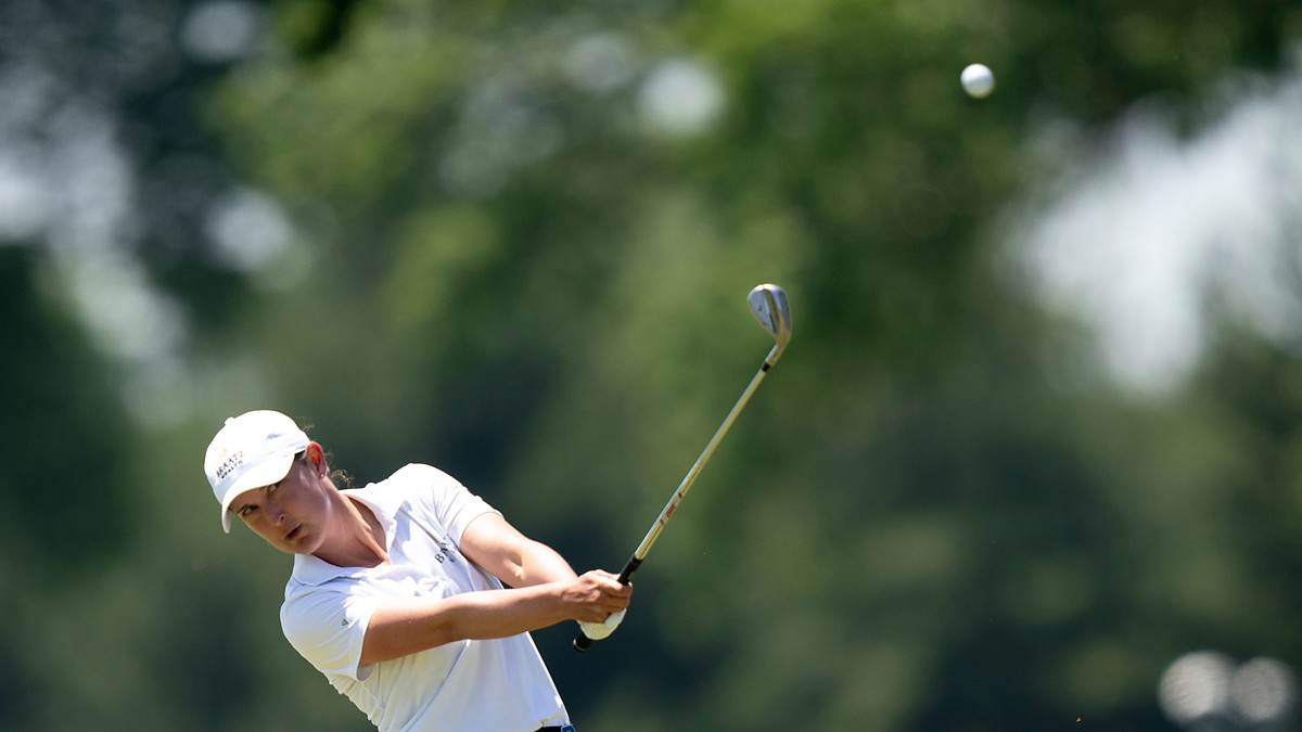 Golfer Lindy Duncan follows through on an iron shot as the ball flies against a backdrop of blurred trees.