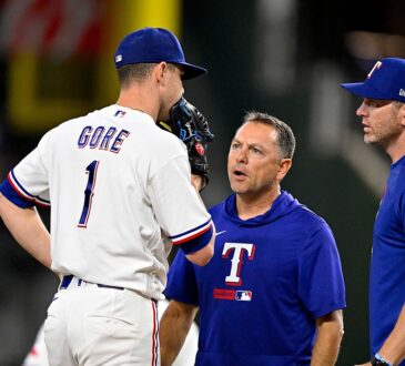 Rangers starting pitcher MacKenzie Gore stands on the mound talking with two members of the coaching and training staff.