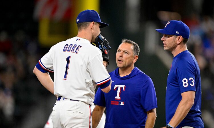 Rangers starting pitcher MacKenzie Gore stands on the mound talking with two members of the coaching and training staff.