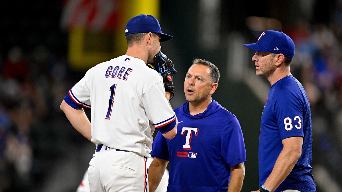 Rangers starting pitcher MacKenzie Gore stands on the mound talking with two members of the coaching and training staff.