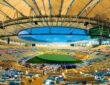 Interior of Maracanã Stadium with an open oval roof, empty yellow and blue seating bowl, and a green field under blue sky.