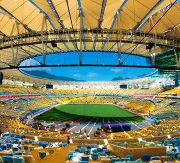 Interior of Maracanã Stadium with an open oval roof, empty yellow and blue seating bowl, and a green field under blue sky.