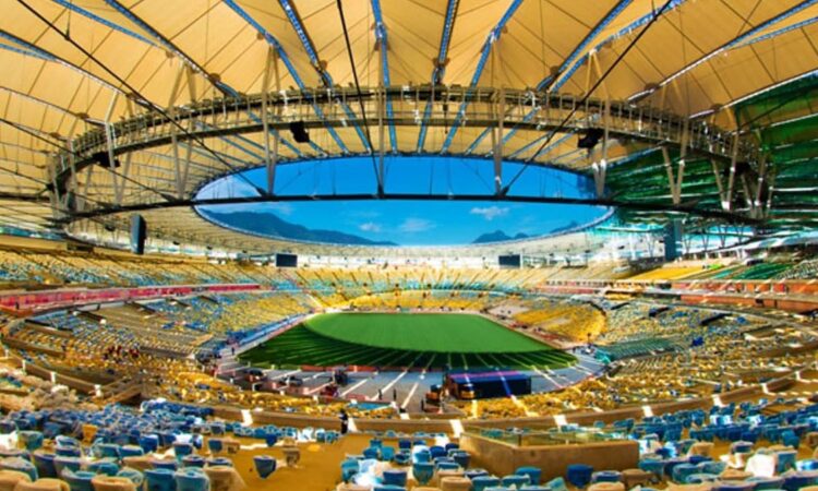 Interior of Maracanã Stadium with an open oval roof, empty yellow and blue seating bowl, and a green field under blue sky.