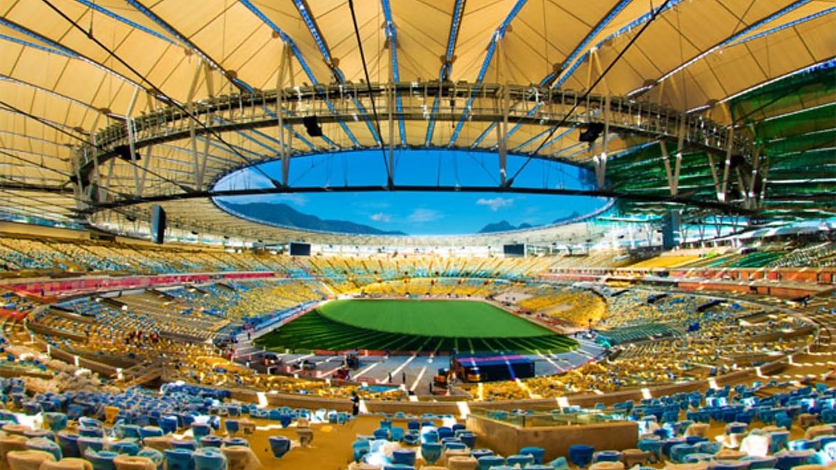Interior of Maracanã Stadium with an open oval roof, empty yellow and blue seating bowl, and a green field under blue sky.
