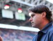 U.S. men’s national team coach Mauricio Pochettino raises his hand inside a stadium as a blurred crowd fills the stands behind him.