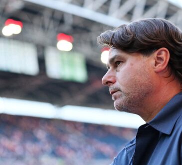 U.S. men’s national team coach Mauricio Pochettino raises his hand inside a stadium as a blurred crowd fills the stands behind him.