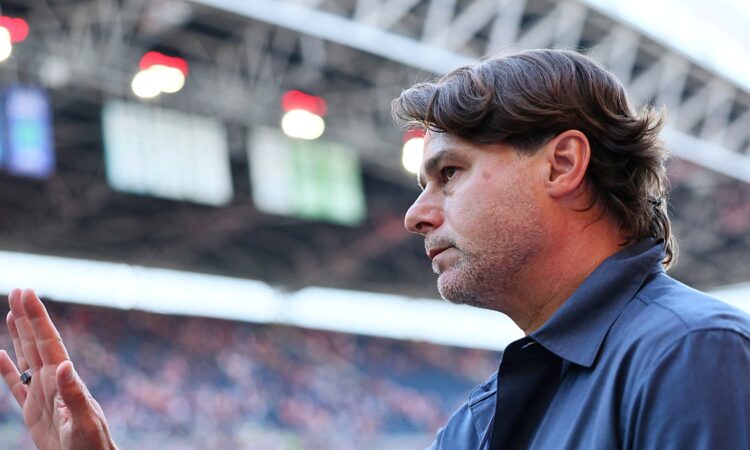 U.S. men’s national team coach Mauricio Pochettino raises his hand inside a stadium as a blurred crowd fills the stands behind him.
