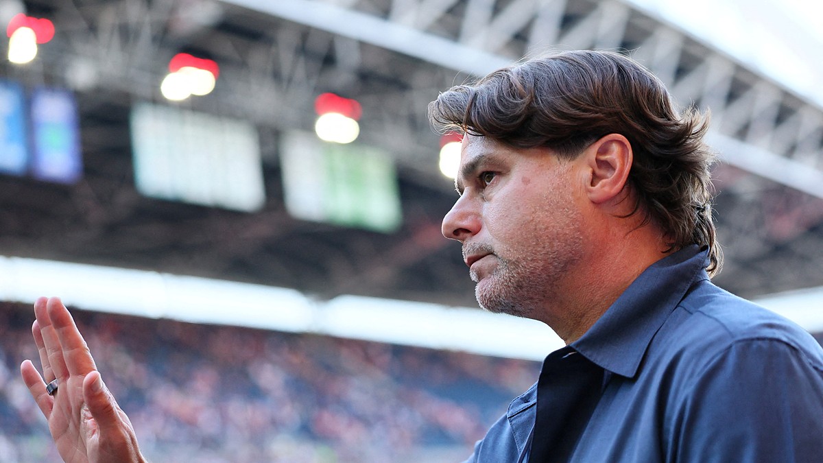 U.S. men’s national team coach Mauricio Pochettino raises his hand inside a stadium as a blurred crowd fills the stands behind him.