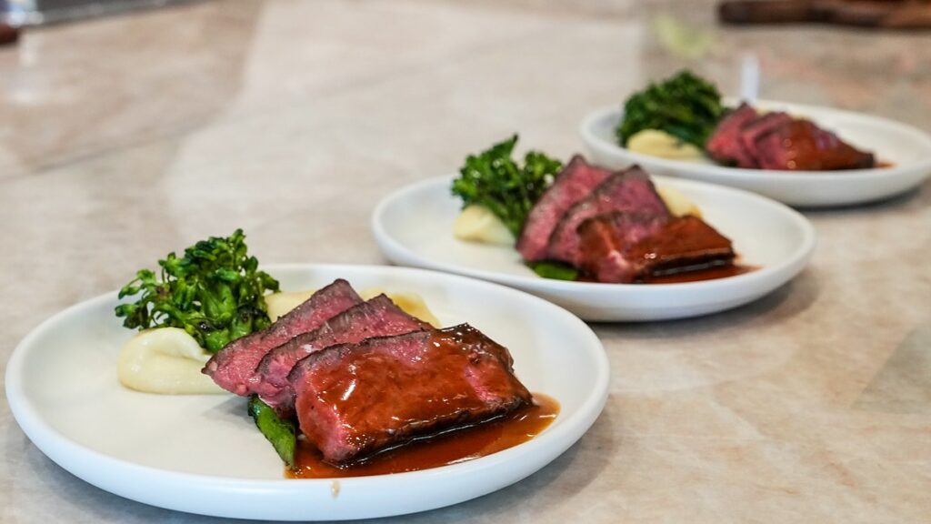 Dallas Mavericks Champions Performance Kitchen plates of sliced steak with sauce, puree, and grilled greens on a marble counter.