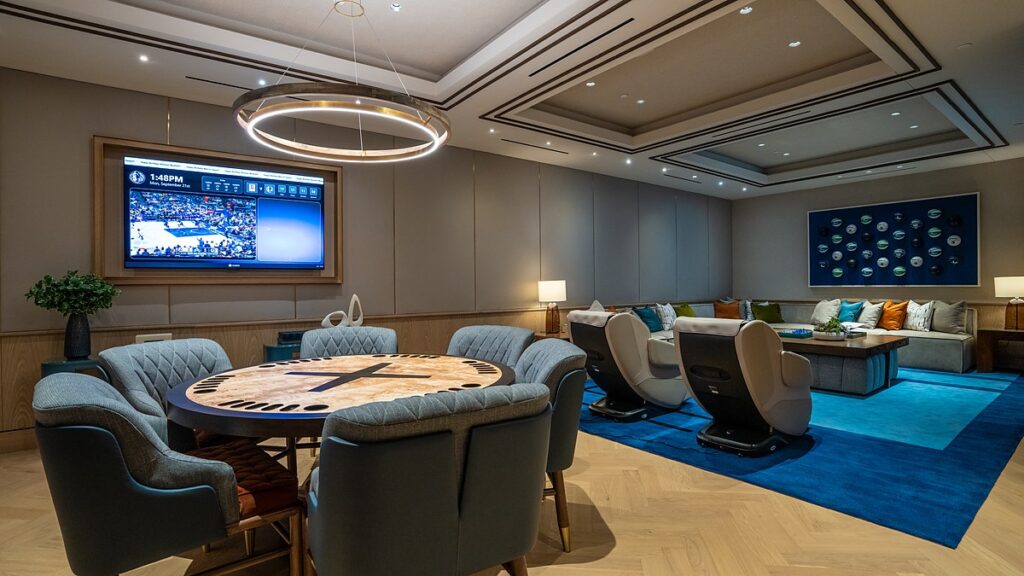 Dallas Mavericks players’ lounge with a round card table, massage chairs, a blue rug, and wall-mounted TVs.