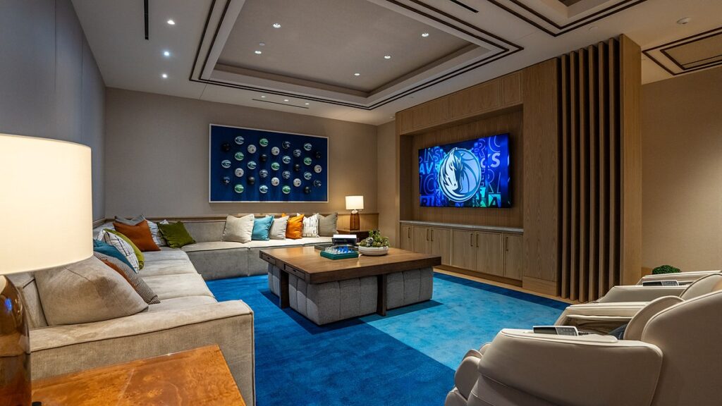 Dallas Mavericks players’ lounge sectional sofa facing a TV showing the Mavericks logo, with massage chairs in the foreground.