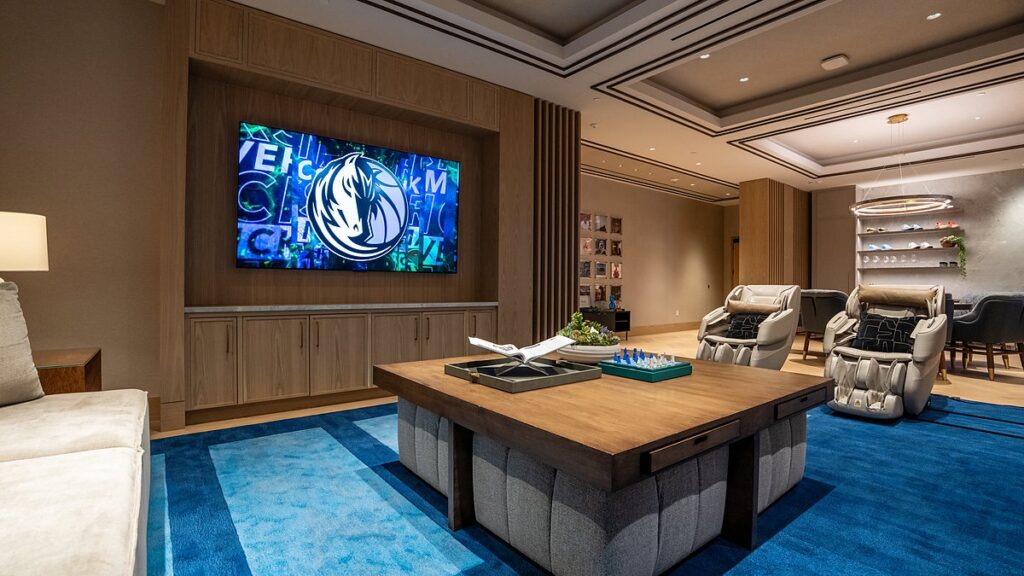 Dallas Mavericks players’ lounge TV displaying the Mavericks logo above a wood coffee table and blue rug.