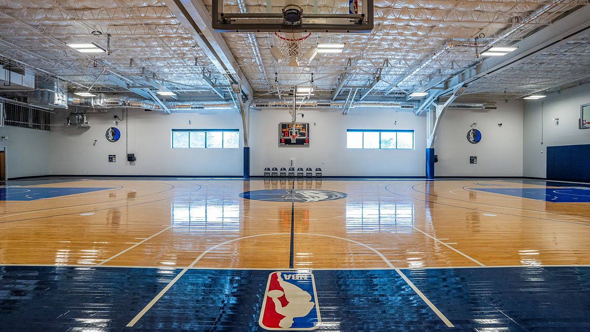 Dallas Mavericks practice court viewed from under a basket, with the NBA logo painted along the baseline.