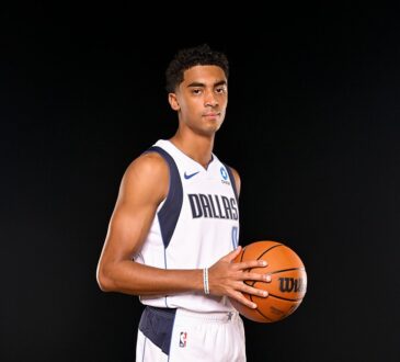 Dallas Mavericks guard Max Christie holds a basketball at his side in a white Dallas jersey against a black backdrop.