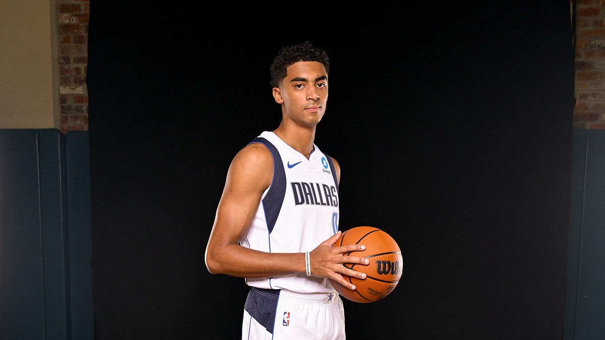 Dallas Mavericks guard Max Christie holds a basketball at his side in a white Dallas jersey against a black backdrop.