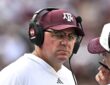 Texas A&M Aggies head coach Mike Elko, wearing a headset and maroon cap, listens to a staff member on the sideline.