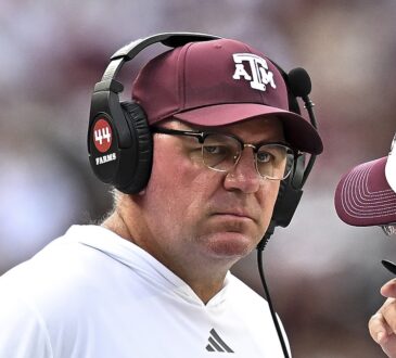 Texas A&M Aggies head coach Mike Elko, wearing a headset and maroon cap, listens to a staff member on the sideline.