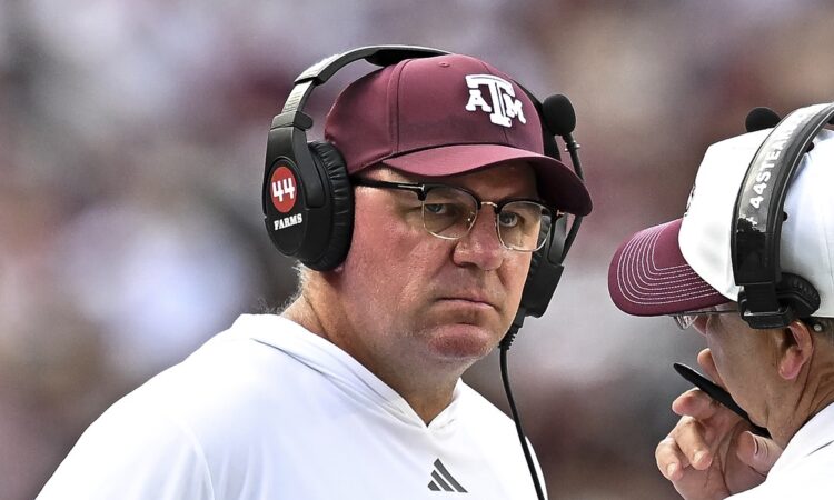 Texas A&M Aggies head coach Mike Elko, wearing a headset and maroon cap, listens to a staff member on the sideline.