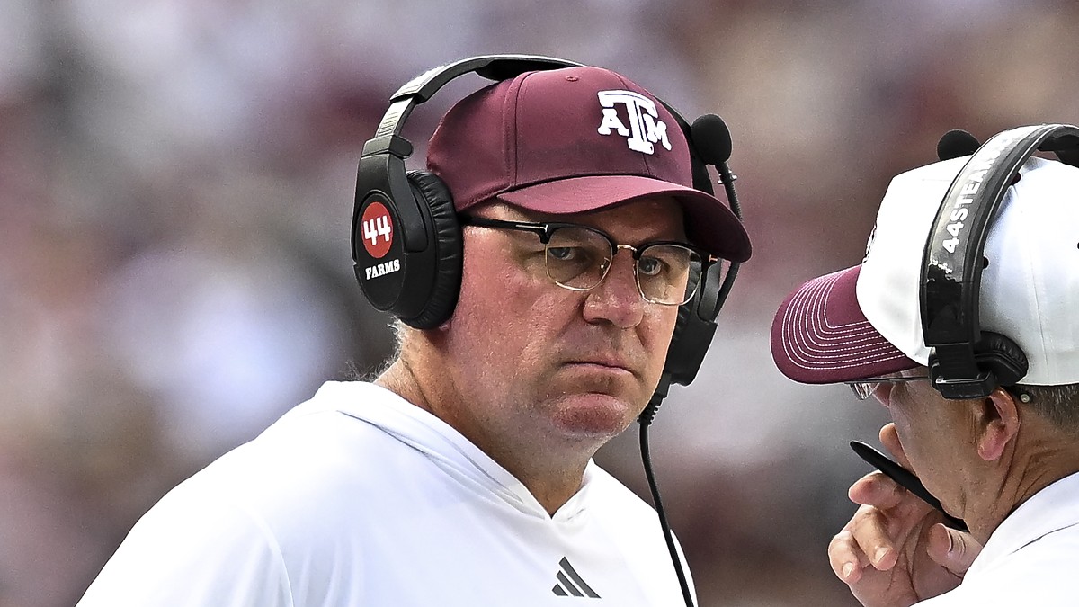 Texas A&M Aggies head coach Mike Elko, wearing a headset and maroon cap, listens to a staff member on the sideline.