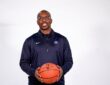 Texas Legends head coach Nathaniel Mitchell smiles and holds a basketball with both hands during a studio portrait.