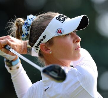 Golfer Nelly Korda watches her tee shot after swinging, wearing a white visor and a blue floral scrunchie.