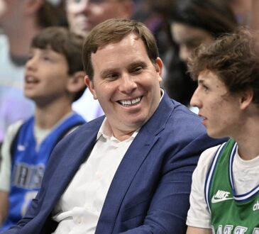Mavericks governor Patrick Dumont smiles in a blue jacket while sitting courtside among fans.