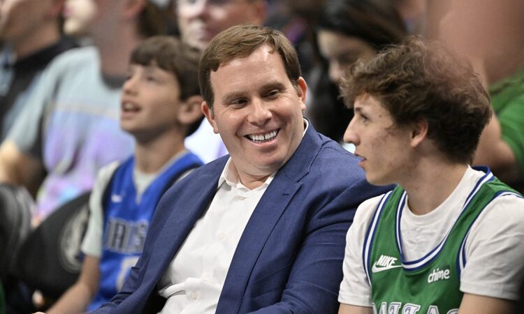 Mavericks governor Patrick Dumont smiles in a blue jacket while sitting courtside among fans.