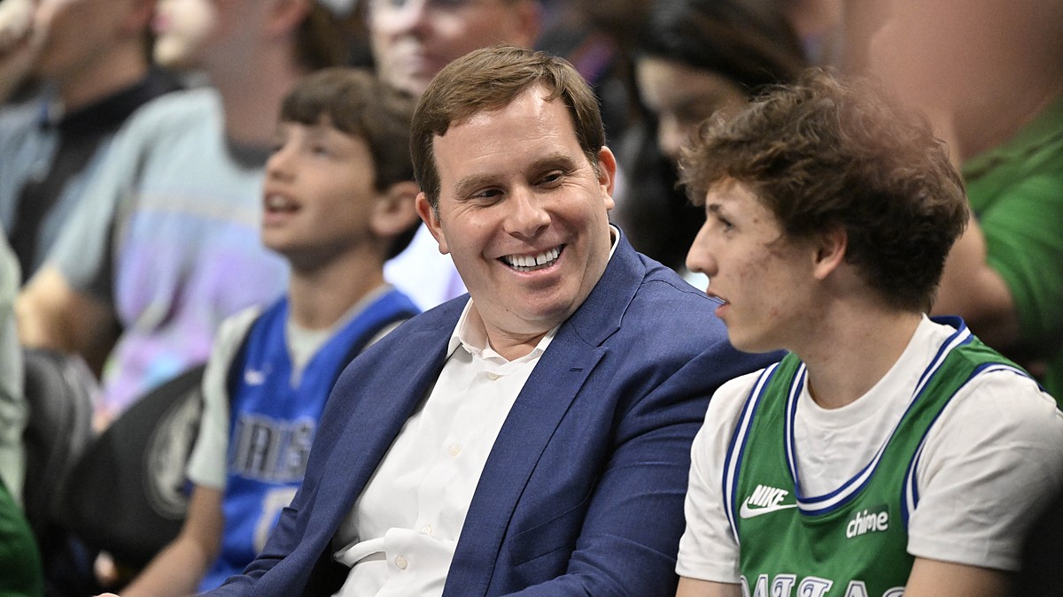 Mavericks governor Patrick Dumont smiles in a blue jacket while sitting courtside among fans.