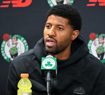 Celtics forward Paul George speaks into a microphone at a podium backed by Celtics logos.