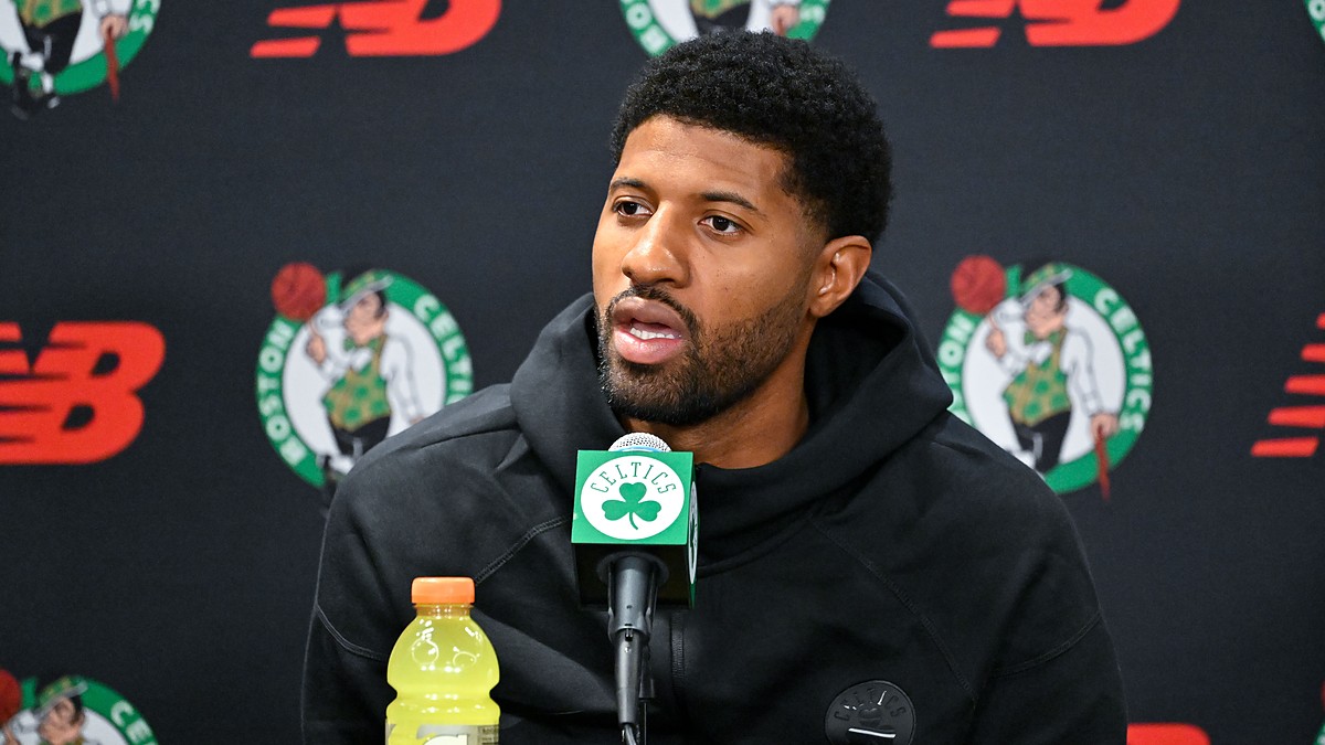 Celtics forward Paul George speaks into a microphone at a podium backed by Celtics logos.