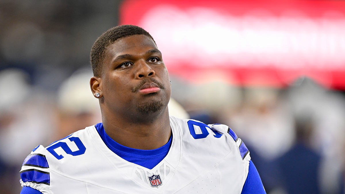 Dallas Cowboys defensive tackle Quinnen Williams gazes upward on the sideline with a blurred video board behind him.