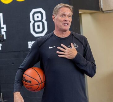 Miami Heat shooting coach Rob Fodor holds a basketball under his arm and speaks with his hand on his chest.