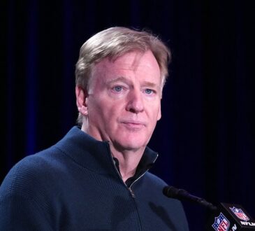 NFL Commissioner Roger Goodell, in a navy quarter-zip sweater, stands at a microphone with an NFL Network mic flag.